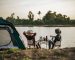 Rear view of young backpacker couple sitting to relax at front of the tent near the lake with coffee set and making fresh coffee grinder while camping trip on summer vacation