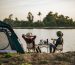 Rear view of young backpacker couple sitting to relax at front of the tent near the lake with coffee set and making fresh coffee grinder while camping trip on summer vacation