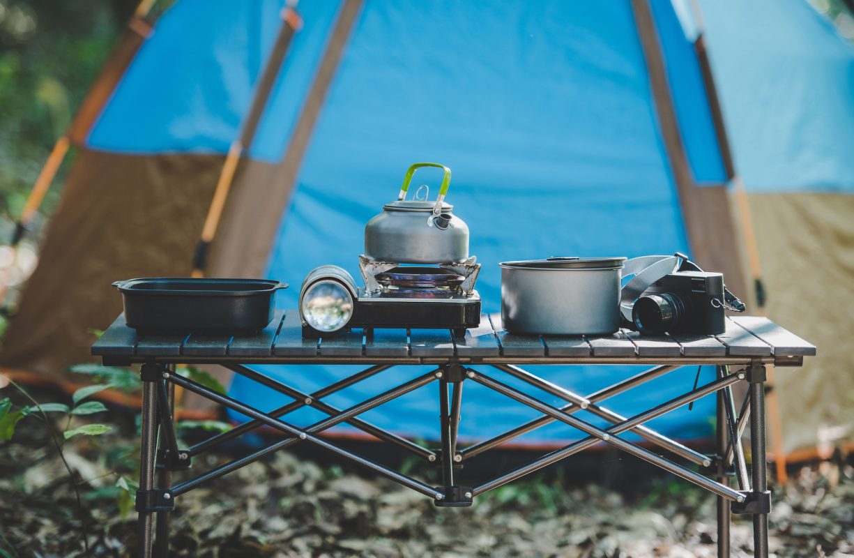 Set of cooking equipment for camping trip with Kettle, pot, pan, flashlight, mini gas stove and camera on table at front of camping tent in nature forest.